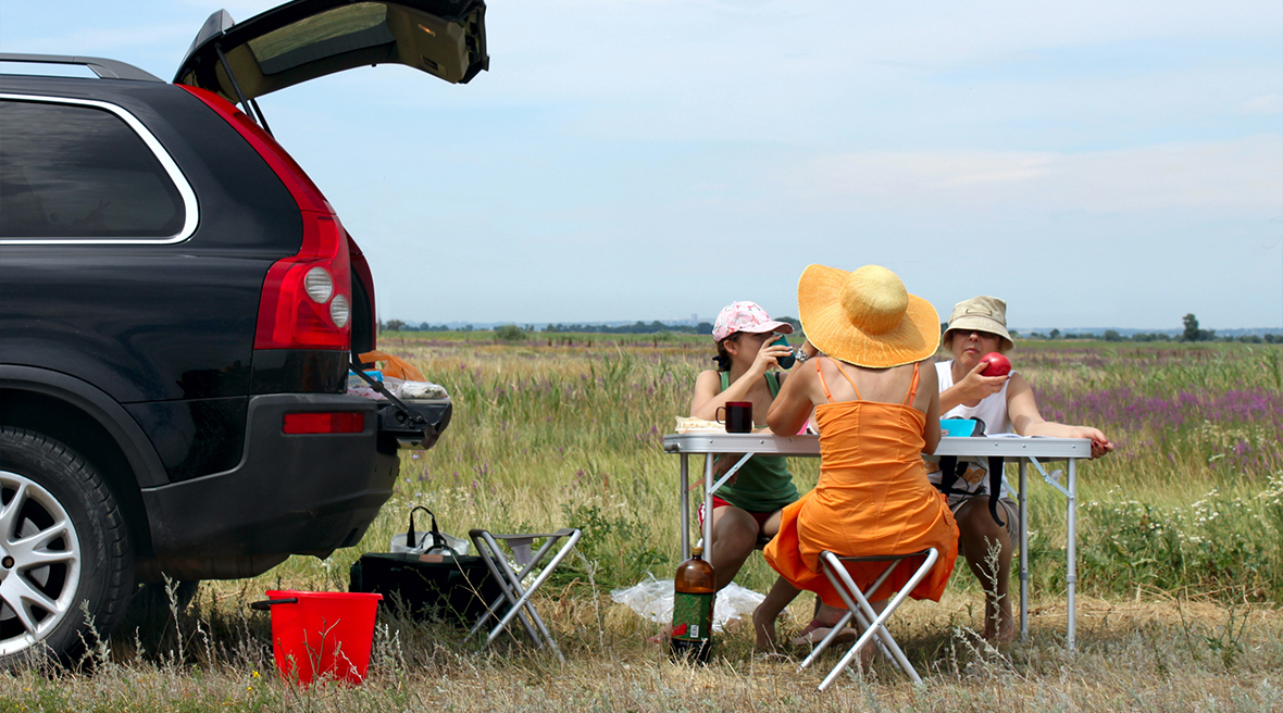 A woman with her back to us in an orange dress and hat with two children opposite her eating at a fold out picnic table from their car which is parked next to them with the boot open